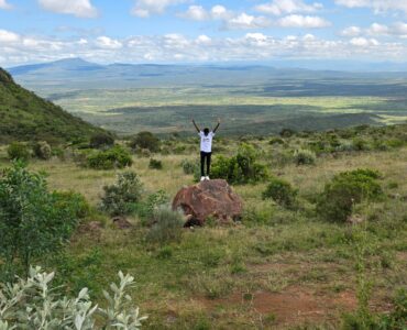 Person celebrating on a rock with arms raised in expansive Kenyan landscape